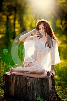 Young beautiful red hair woman wearing a transparent white blouse posing on a stump in a green forest. Fashionable girl