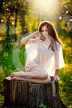 Young beautiful red hair woman wearing a transparent white blouse posing on a stump in a green forest. Fashionable girl