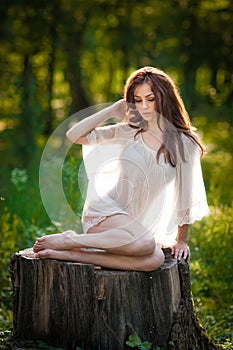 Young beautiful red hair woman wearing a transparent white blouse posing on a stump in a green forest. Fashionable girl