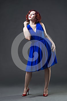 Young beautiful red-haired caucasian woman in blue dress posing in studio on gray background, professional makeup and hairstyle