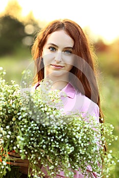 A young beautiful red-haired girl close-up is looking at the camera with a huge bouquet of field daisies in a pink dress. Sunset