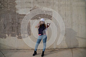 Young and beautiful red-haired woman with baseball cap, jacket with baseball bat defying camera. she is on grey concrete