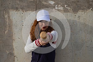 Young and beautiful red-haired woman with baseball cap, jacket with baseball bat defying camera. she is on grey concrete