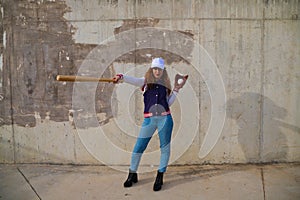 Young and beautiful red-haired woman with baseball cap, jacket with baseball bat defying camera. she is on grey concrete