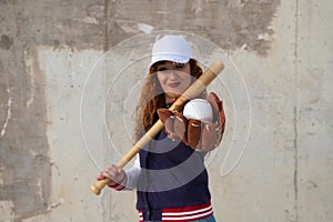 Young and beautiful red-haired woman with baseball cap, jacket and glove with baseball bat resting on her shoulder on grey cement