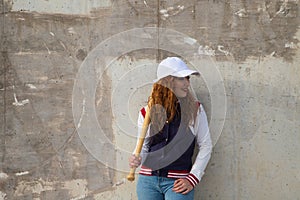 Young and beautiful red-haired woman with baseball cap, jacket and glove with baseball bat resting on her shoulder on grey cement