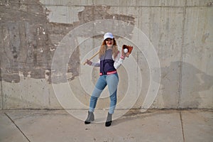 Young and beautiful red-haired woman with baseball cap, jacket and glove with baseball bat resting on her shoulder on grey cement