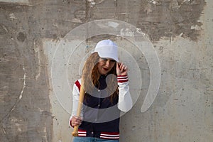 Young and beautiful red-haired woman with baseball cap, jacket and glove with baseball bat resting on her shoulder on grey cement