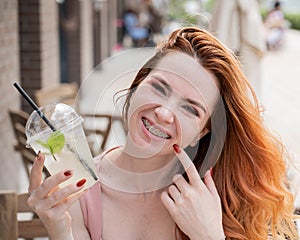 Young beautiful red-haired woman with braces drinks cooling lemonade outdoors in summer. Portrait of a smiling girl with