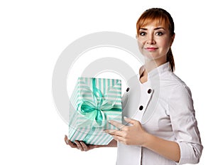 Young beautiful red-haired woman doctor or nurse in white special uniform standing and holding big green present box with ribbon