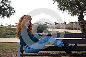 Young and beautiful red-haired woman is sitting on a bench in a park where there are many olive trees. She is on her laptop