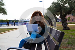 Young and beautiful red-haired woman is sitting on a bench in a park where there are many olive trees. She is on her laptop