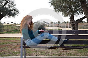 Young and beautiful red-haired woman is sitting on a bench in a park where there are many olive trees. She is on her laptop