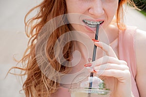 Young beautiful red-haired woman smiling with braces drinks cooling lemonade outdoors in summer. Cropped portrait Girl