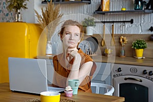 Young beautiful red-haired woman using a laptop computer and drinking a morning cup of coffee or tea. He looks thoughtfully out