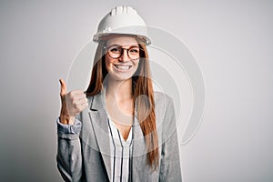 Young beautiful redhead architect woman wearing security helmet over white background doing happy thumbs up gesture with hand