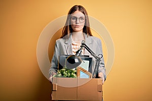 Young beautiful redhead businesswoman fired holding carboard box over yellow background with a confident expression on smart face