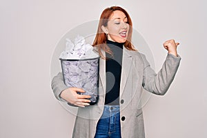 Young beautiful redhead businesswoman holding full paper bin with crumpled papers pointing and showing with thumb up to the side