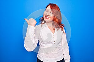 Young beautiful redhead businesswoman wearing elegant shirt standing over blue background pointing thumb up to the side smiling