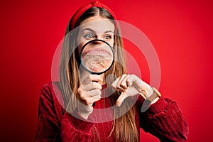 Young beautiful redhead detective woman using magnifying glass over isolated red background with angry face, negative sign showing