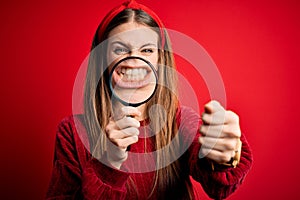 Young beautiful redhead detective woman using magnifying glass over isolated red background annoyed and frustrated shouting with