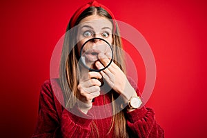 Young beautiful redhead detective woman using magnifying glass over isolated red background cover mouth with hand shocked with