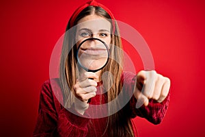 Young beautiful redhead detective woman using magnifying glass over isolated red background pointing with finger to the camera and