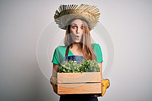 Young beautiful redhead farmer woman wearing apron and hat holding box with plants scared in shock with a surprise face, afraid