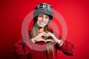 Young beautiful redhead motocyclist woman wearing moto helmet over red background smiling in love showing heart symbol and shape