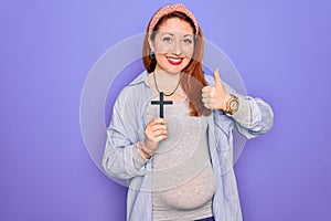 Young beautiful redhead pregnant woman expecting baby holding christian cross happy with big smile doing ok sign, thumb up with