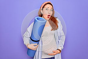 Young beautiful redhead pregnant woman expecting baby holding mat to do prenatal exercise scared in shock with a surprise face,