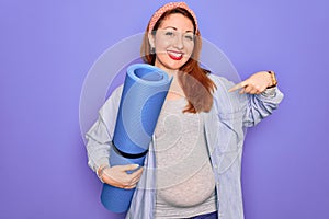 Young beautiful redhead pregnant woman expecting baby holding mat to do prenatal exercise with surprise face pointing finger to