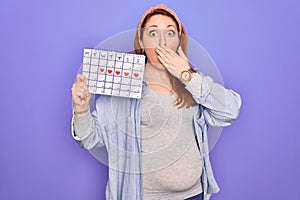 Young beautiful redhead pregnant woman expecting baby holding period calendar cover mouth with hand shocked with shame for
