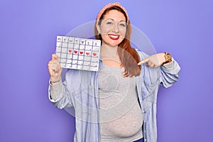 Young beautiful redhead pregnant woman expecting baby holding period calendar with surprise face pointing finger to himself
