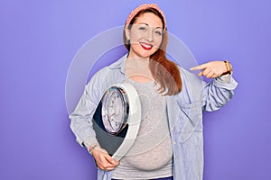 Young beautiful redhead pregnant woman expecting baby holding weighting machine with surprise face pointing finger to himself