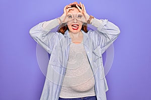 Young beautiful redhead pregnant woman expecting baby over isolated purple background doing ok gesture like binoculars sticking