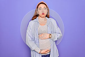 Young beautiful redhead pregnant woman expecting baby over isolated purple background puffing cheeks with funny face