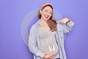 Young beautiful redhead pregnant woman expecting baby with question mark on stomach with surprise face pointing finger to himself