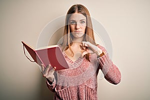Young beautiful redhead student woman reading book over isolatated white background with angry face, negative sign showing dislike