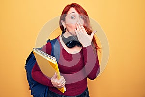 Young beautiful redhead student woman wearing backpack and headphones holding notebook covering mouth with hand, shocked and