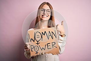 Young beautiful redhead woman asking for women rights holding banner over pink background happy with big smile doing ok sign,