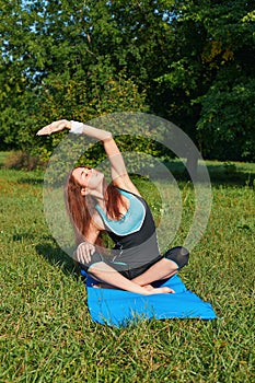 Young beautiful redhead woman doing yoga on a yoga mat. Performing yoga asanas in a green park. Sport, fitness and lifestyle