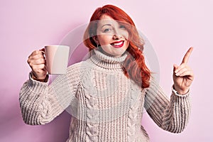 Young beautiful redhead woman drinking cup of coffee over isolated pink background smiling happy pointing with hand and finger to