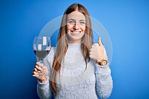 Young beautiful redhead woman drinking glass of red wine over isolated blue background happy with big smile doing ok sign, thumb