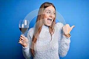 Young beautiful redhead woman drinking glass of red wine over isolated blue background pointing and showing with thumb up to the
