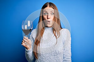 Young beautiful redhead woman drinking glass of red wine over isolated blue background scared in shock with a surprise face,