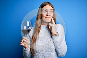 Young beautiful redhead woman drinking glass of red wine over isolated blue background serious face thinking about question, very