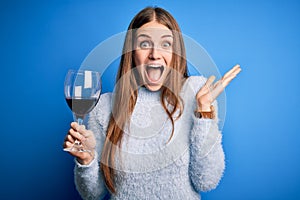 Young beautiful redhead woman drinking glass of red wine over isolated blue background very happy and excited, winner expression