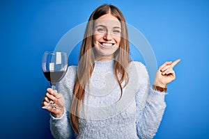 Young beautiful redhead woman drinking glass of red wine over isolated blue background very happy pointing with hand and finger to