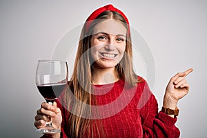 Young beautiful redhead woman drinking glass of red wine over isolated white background very happy pointing with hand and finger